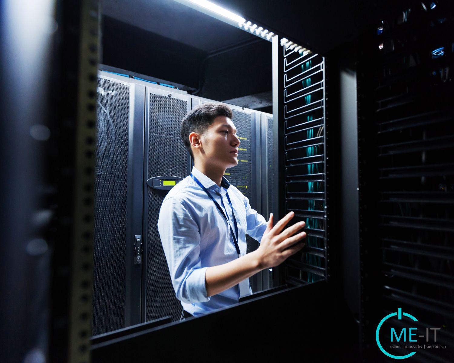 A technician is working inside a server room, inspecting and managing server equipment placed on racks. The person is wearing formal attire with an ID badge, and is surrounded by network cables and server cabinets. The setting is modern, with visible LED lighting and the ME-IT company logo in the lower right corner.